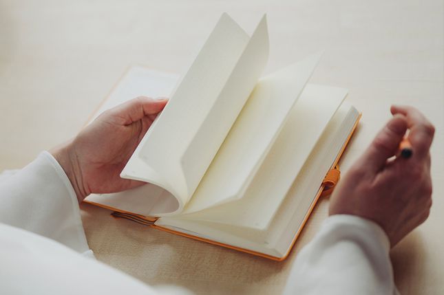 Verena Sons Eine Frau blättert in einem Buch mit leeren Seiten und hält einen orangen Stift in der Hand.